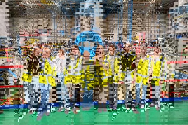 Een groep van tien jonge meiden met gele veiligheidsvesten en vrijetijdskleding staat op een rij in een industriële omgeving met een KLM vliegtuig op de achtergrond.