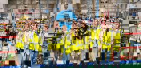 Een groep van tien jonge meiden met gele veiligheidsvesten en vrijetijdskleding staat op een rij in een industriële omgeving met een KLM vliegtuig op de achtergrond.