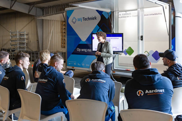 Trainer Wij Techniek Vakbus spreekt tot een groep zittende mensen in een workshopomgeving, met een presentatiescherm erachter.