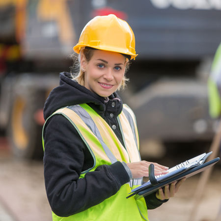 role="presentation" Vrouwelijke technisch medewerker op bouwplaats met gele helm, geel jasje en ordner in de hand, kijkt lachend in de camera.