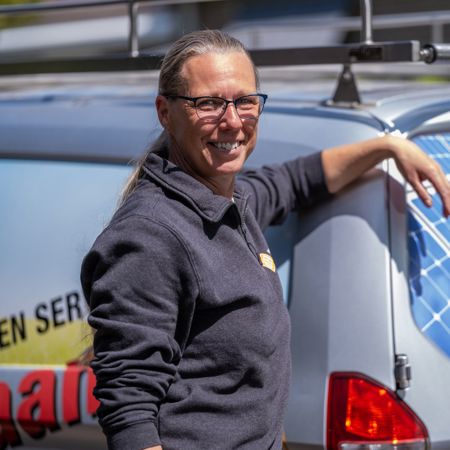 Vrouwelijke installateur kijkt lachend naar de camera en leunt tegen achterkant van werkbus met daarop logo van Installatiebedrijf De Haan.