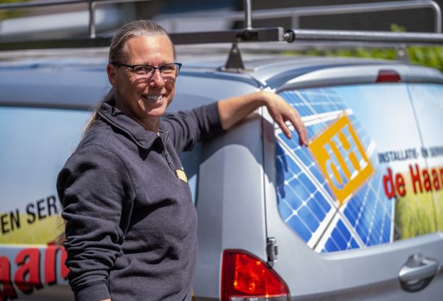 Vrouwelijke installateur kijkt lachend naar de camera en leunt tegen achterkant van werkbus met daarop logo van Installatiebedrijf De Haan.