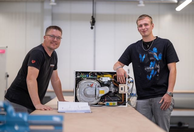 Twee jonge monteurs van Lomans Academy leunend op een werkbank, kijken lachend naar de camera, terwijl ze bezig zijn met installatie.