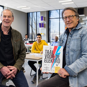 De twee schrijvers van Taal voor de elektro kijken lachend en trost in de camera. Ze zitten beiden op een tafel in een klaslokaal. Eén van hen heeft het boek in de hand. 