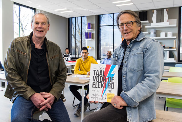 De twee schrijvers van Taal voor de elektro kijken lachend en trost in de camera. Ze zitten beiden op een tafel in een klaslokaal. Eén van hen heeft het boek in de hand.